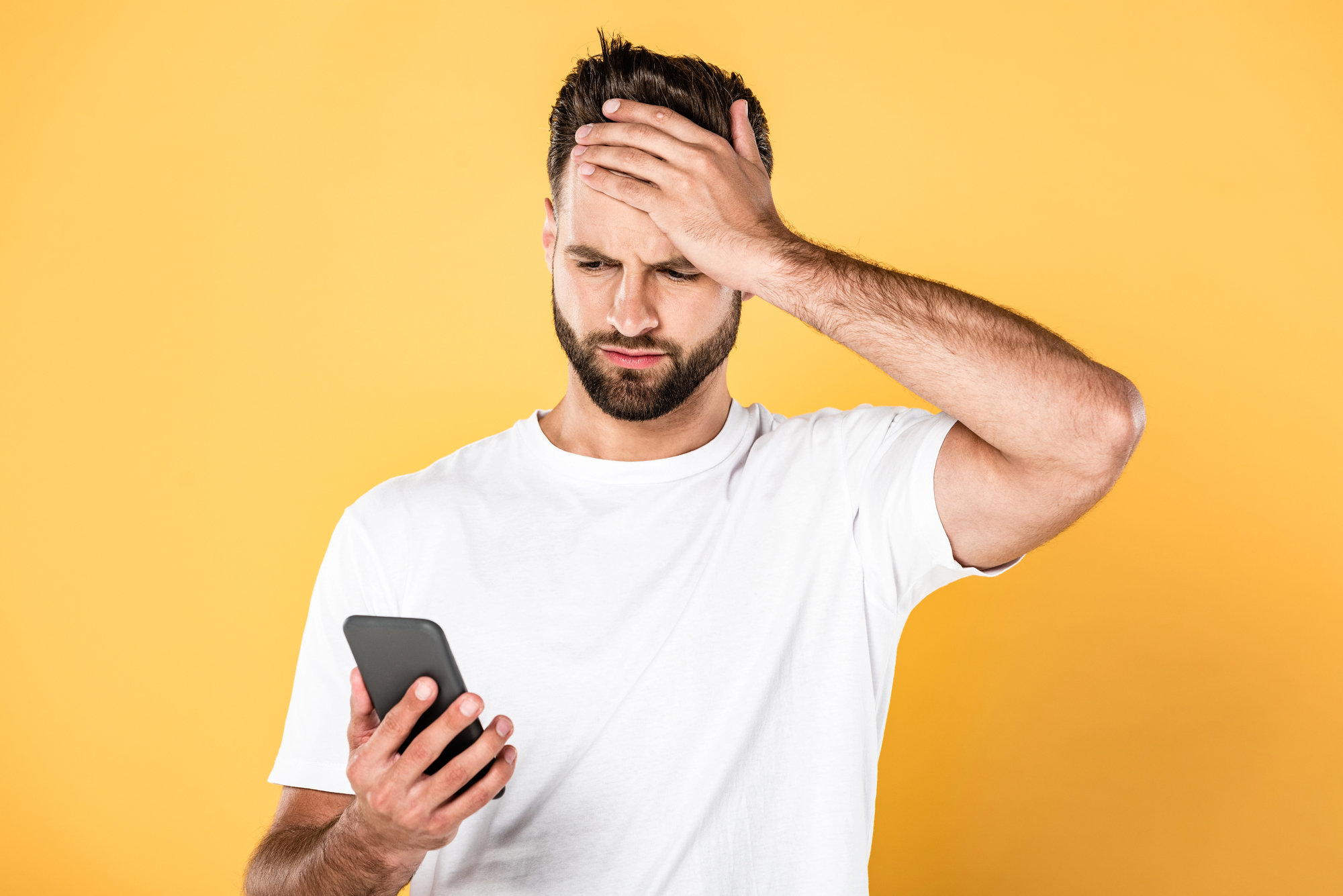 Upset man in white t-shirt with facepalm using smartphone isolated on yellow
