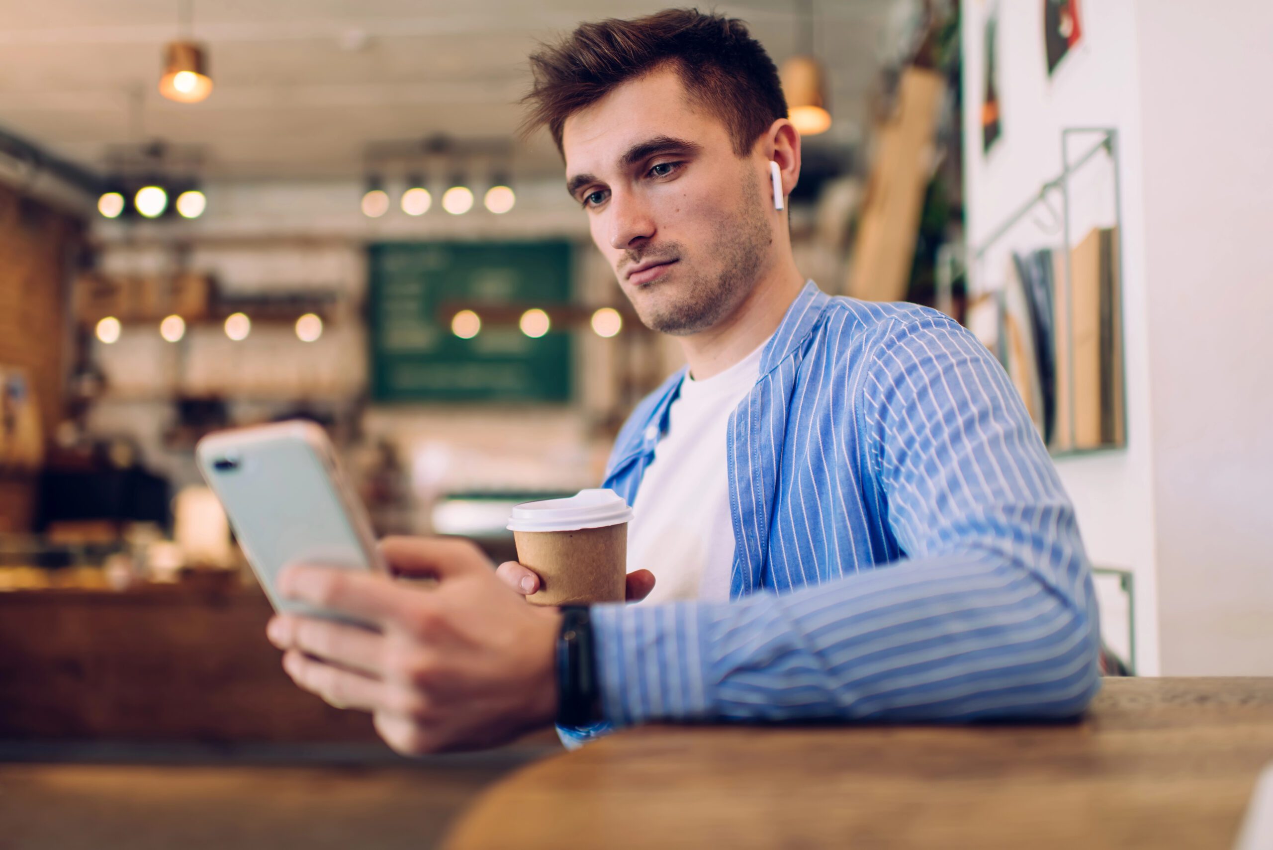 Back view of serious man with dark hair in casual clothes holding coffee and browsing smartphone while messaging with friends in cafeteria