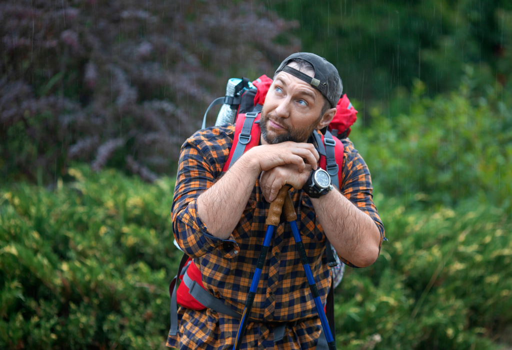 A man with a backpack rests on his trekking poles in a green forest.